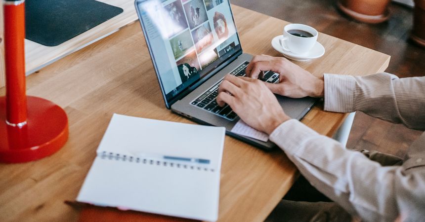 Hombre escribe en un computador en su hogar, acompañado de una taza de café y de un cuaderno de apuntes abierto sobre la mesa.