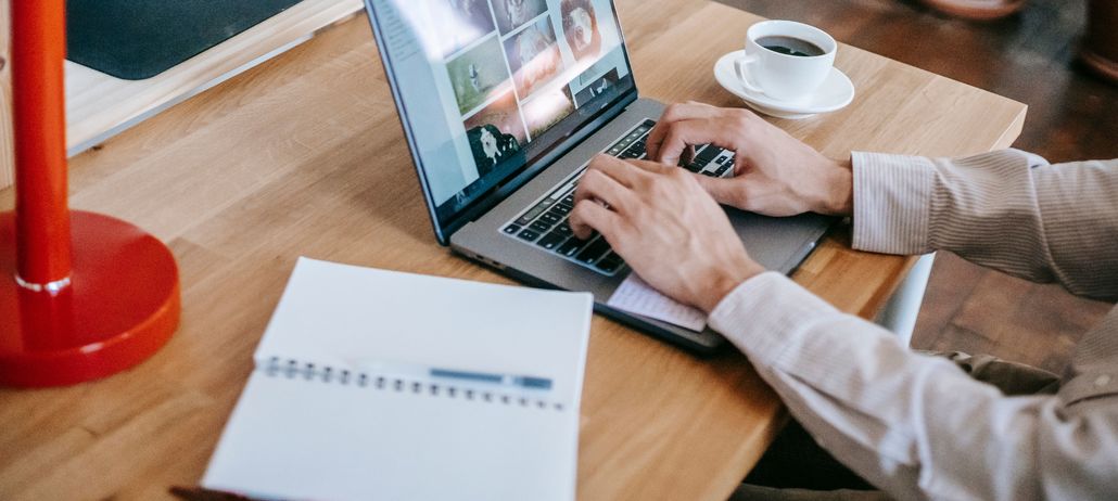 Hombre escribe en un computador en su hogar, acompañado de una taza de café y de un cuaderno de apuntes abierto sobre la mesa.